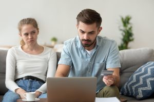 Couple sitting in living room at home. Unsatisfied wife looking at husband, man holding credit card choose goods on internet online, using laptop banking payment services problems, fraud scam concept