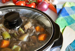 crock pot with beef stew in it and a checked blue and green cloth on the table and tomatoes and onions in the top left on the table