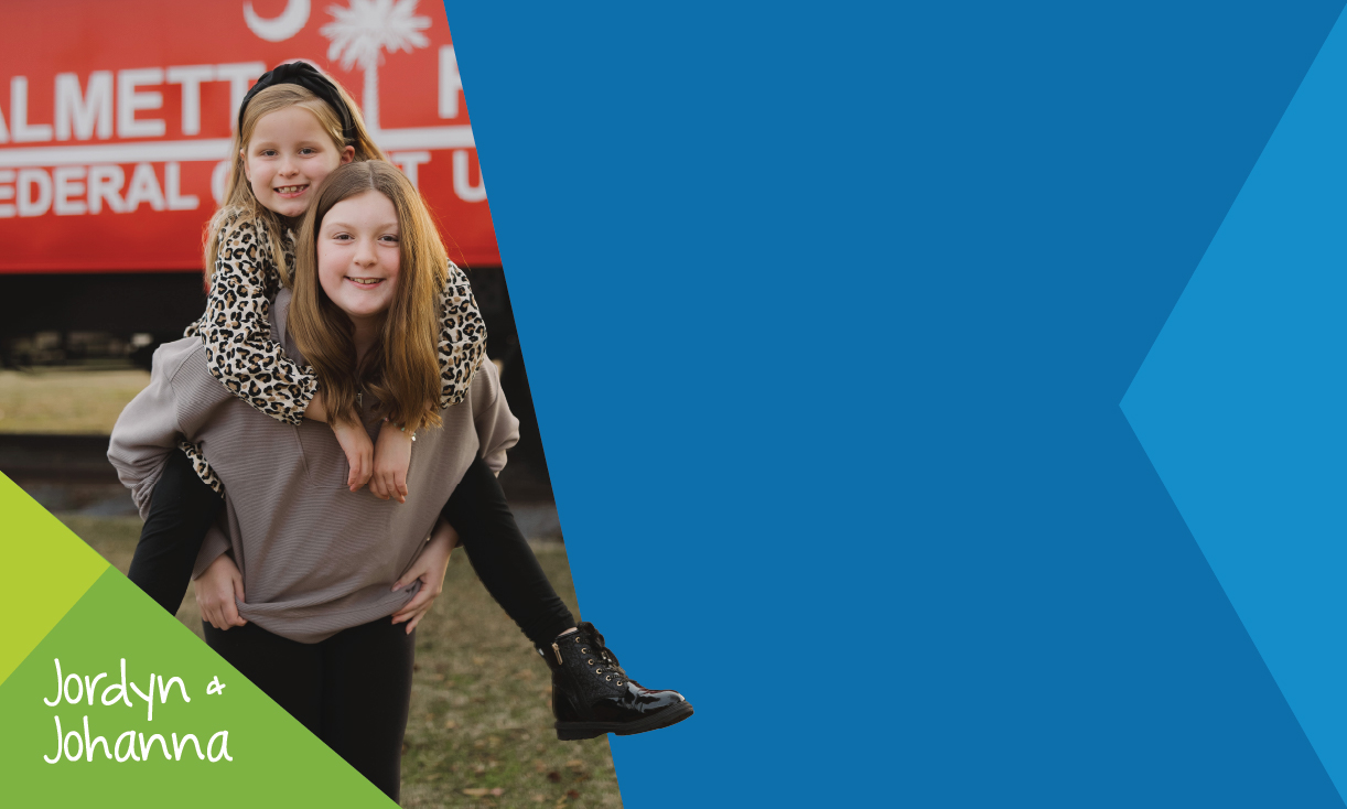 Two smiling girls, one giving a piggyback ride, in front of a red train. They wear casual clothes. Text says "Jordyn & Johanna" on a green triangle.