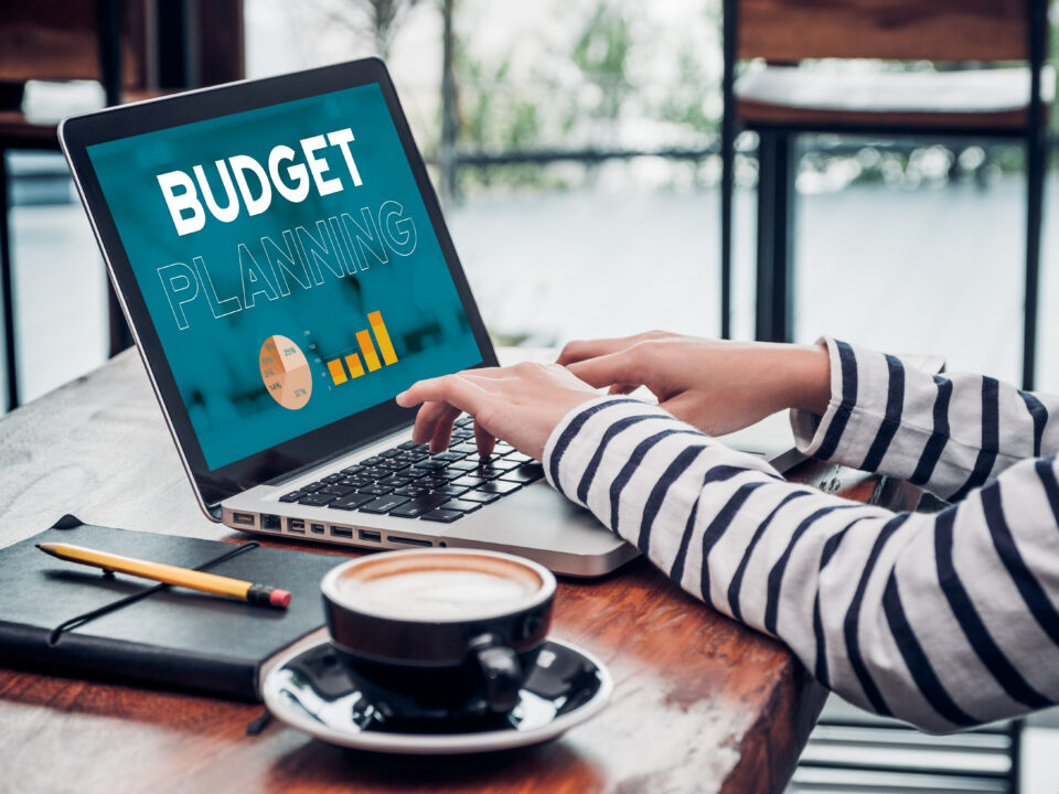 A person in a striped shirt is typing on a laptop displaying "Budget Planning" with charts. A notebook, pencil, and coffee cup are on the table.