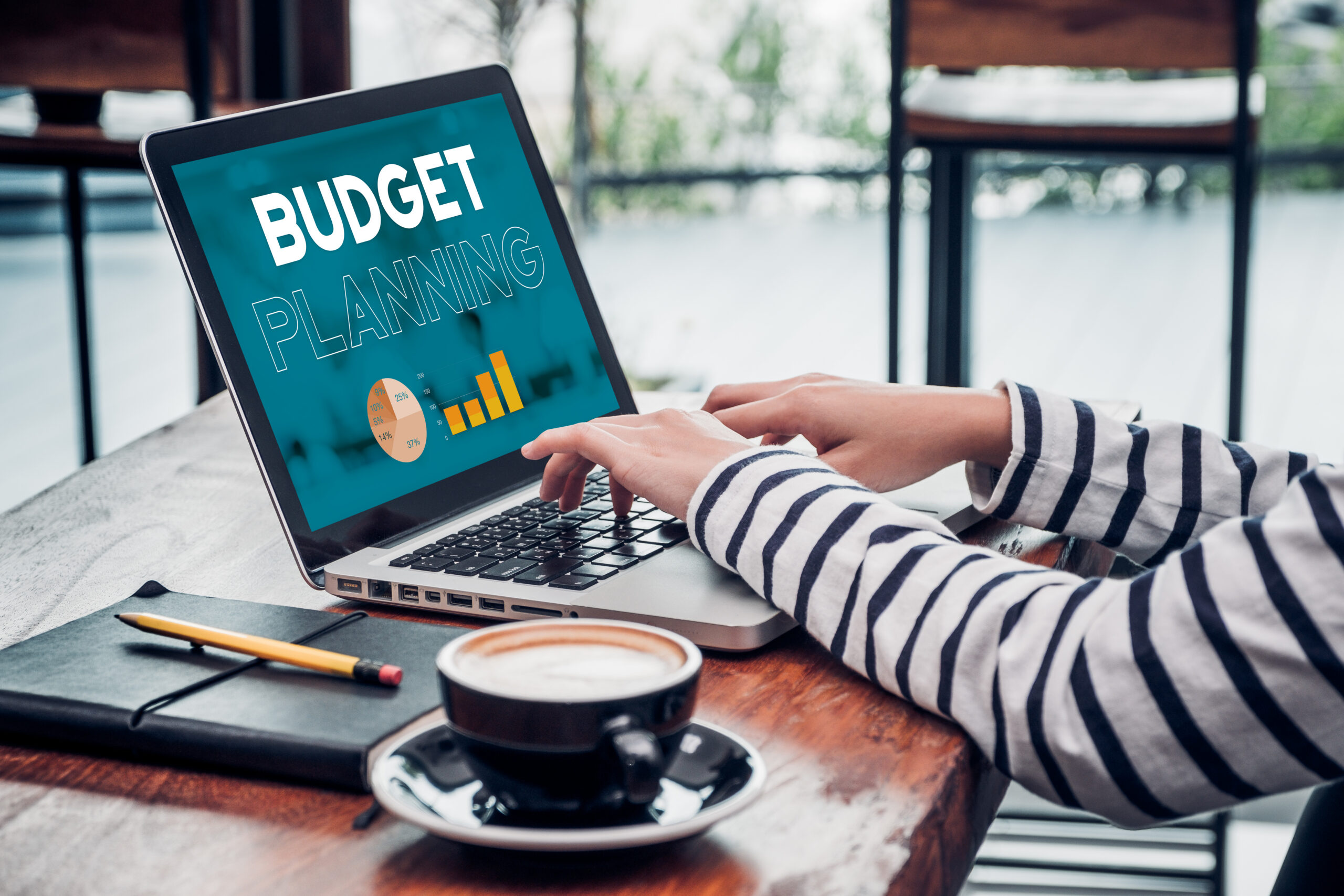 A person in a striped shirt is typing on a laptop displaying "Budget Planning" with charts. A notebook, pencil, and coffee cup are on the table.