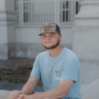 _I5A8823 Young man with a beard and camouflage hat smiles while sitting outdoors