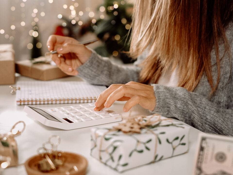 Person using a calculator to plan a holiday gift budget, surrounded by wrapped presents and cash