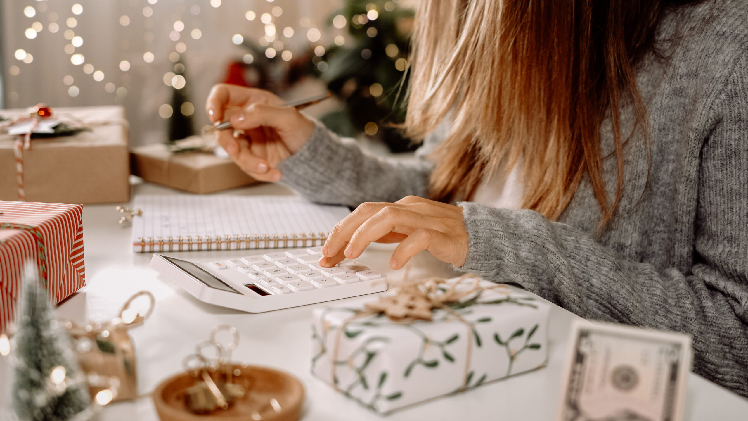 Person using a calculator to plan a holiday gift budget, surrounded by wrapped presents and cash